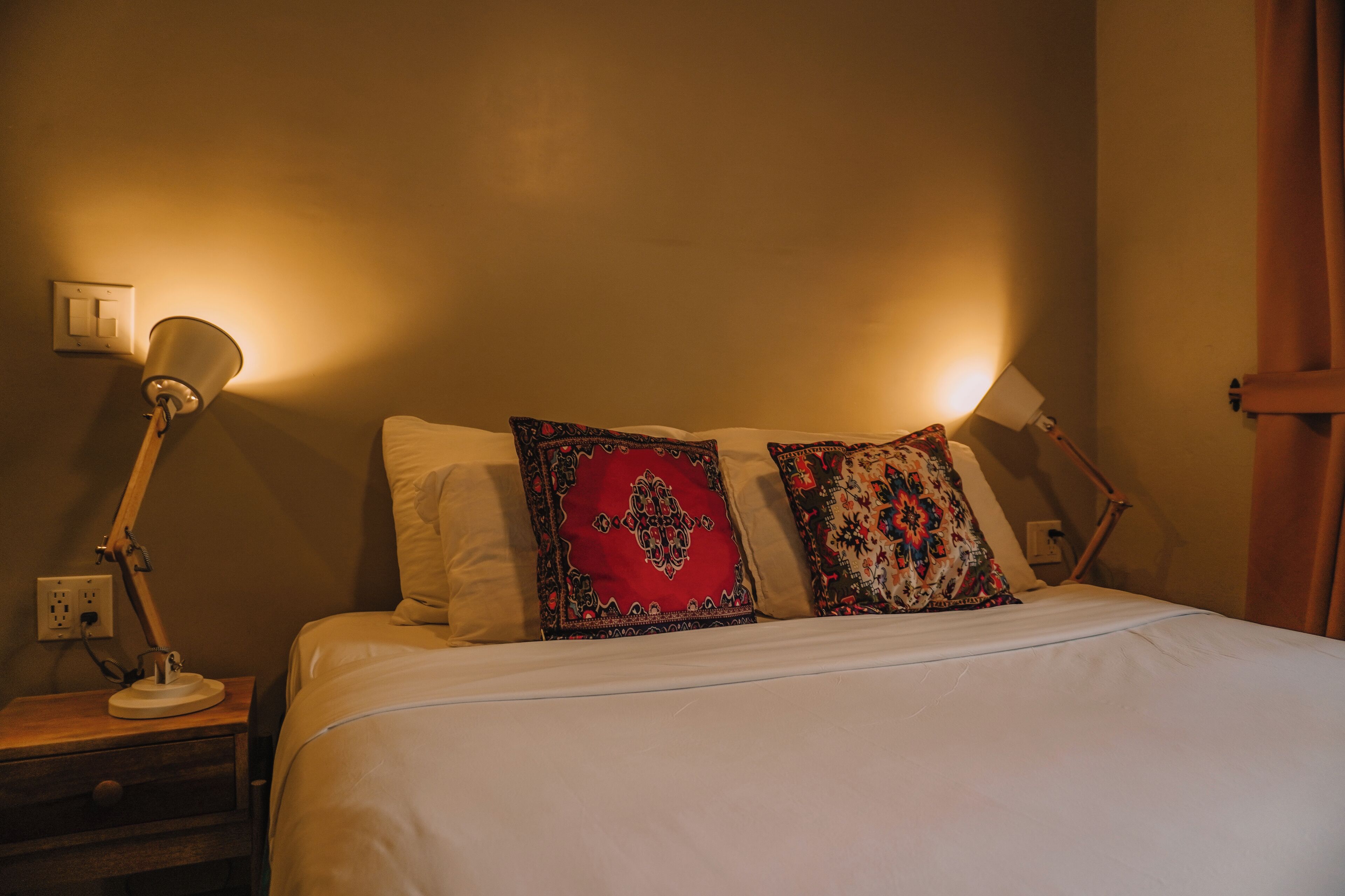 A neatly made bed with white bedding and two decorative pillows, a bedside lamp, and a nightstand with a drawer.