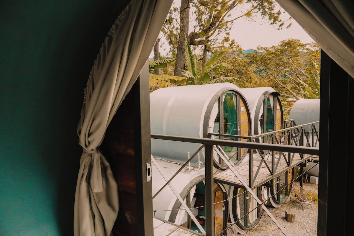 A view through a window with curtains, revealing a series of cylindrical structures with windows, possibly a unique accommodation style, surrounded by trees and a clear sky.