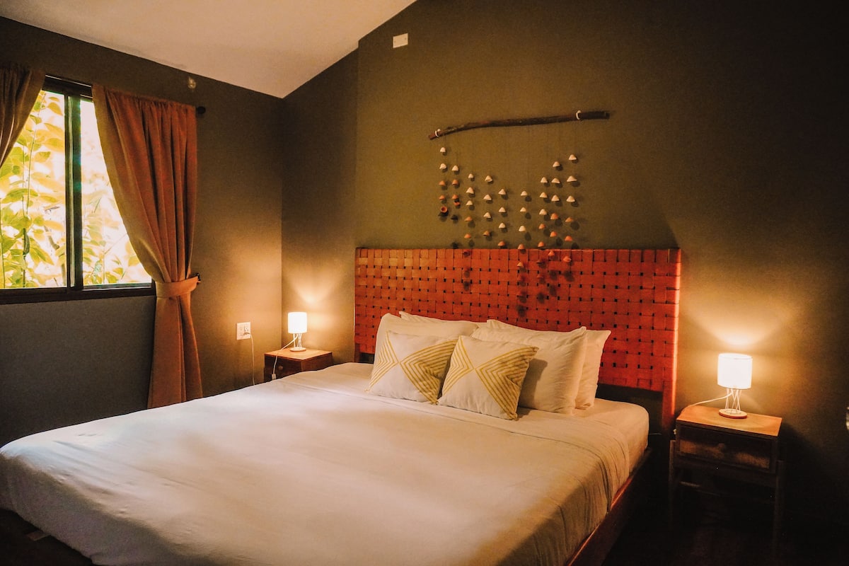 A hotel room with a bed, two bedside tables, a wall-mounted light fixture, and a window with curtains.