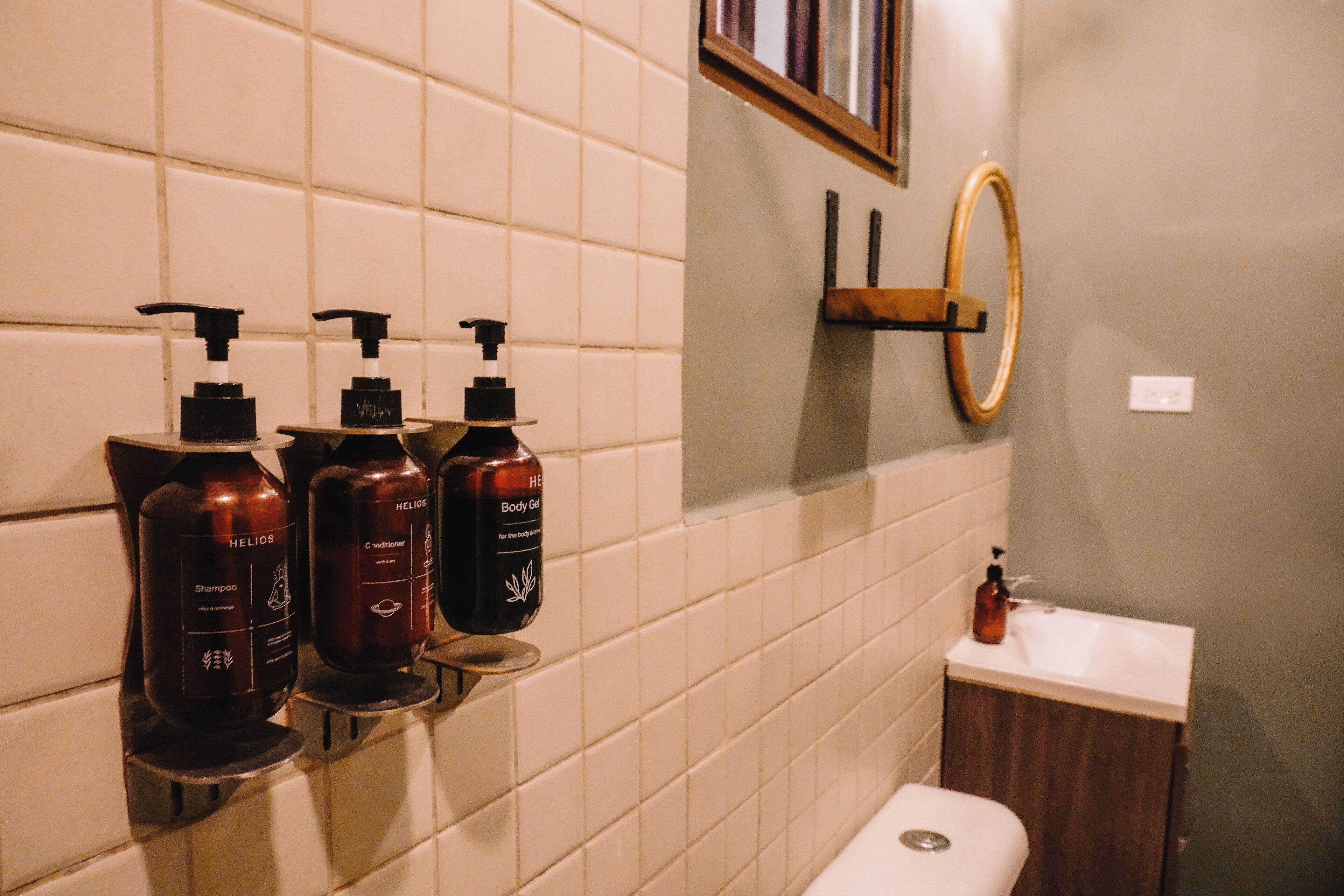 A bathroom with a toilet, a sink, and three bottles of shampoo and conditioner on a wall-mounted shelf.