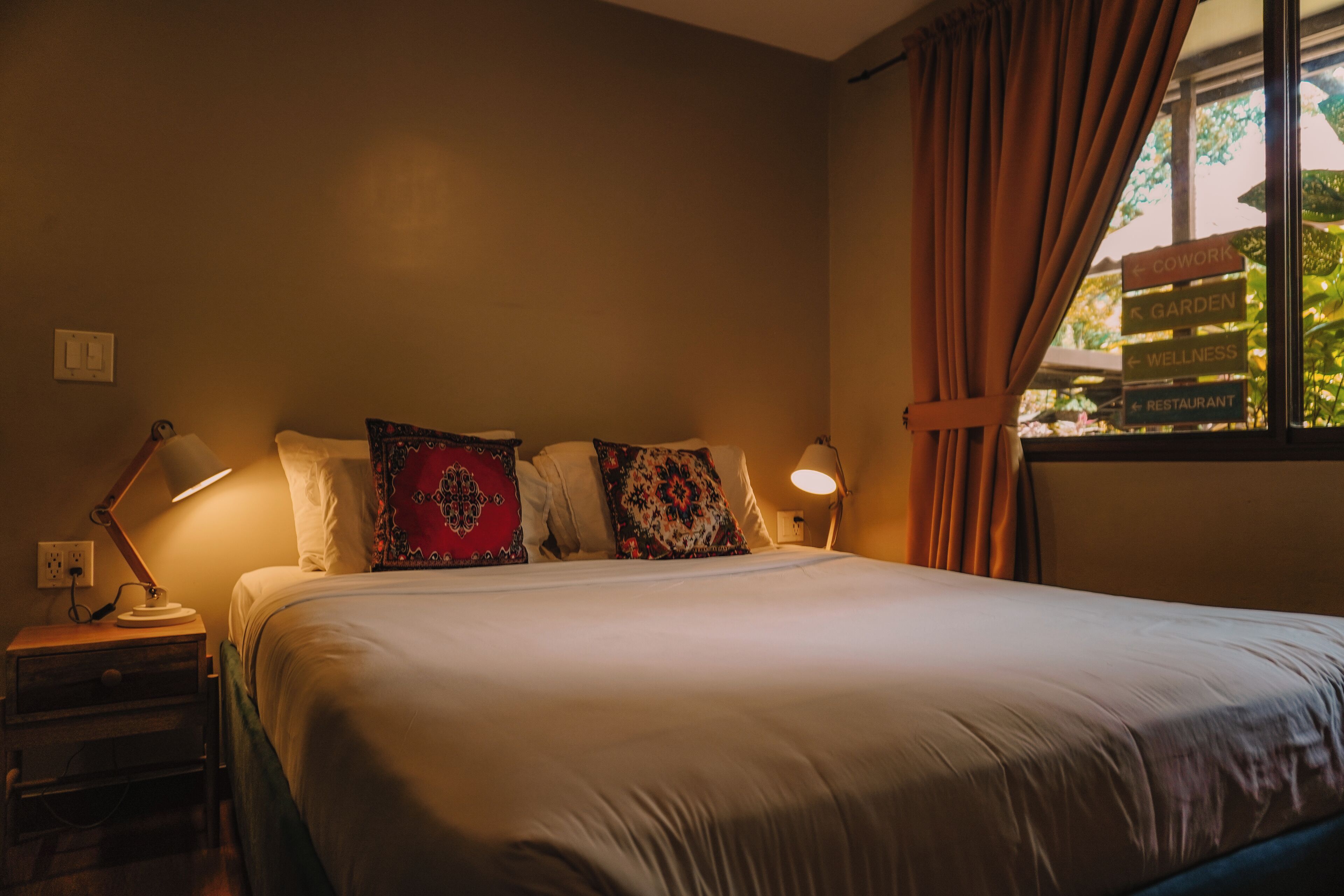 Una cama bien hecha con edredón blanco y varios cojines decorativos. Hay una lámpara de noche sobre una mesita de madera a la izquierda de la cama. La habitación tiene una ventana con cortinas corridas lateralmente, dejando entrar luz natural.