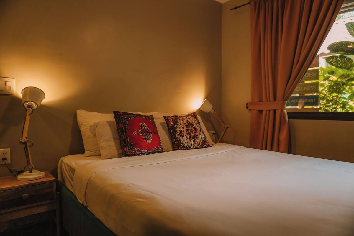 A neatly made bed with white linens and patterned pillows, a bedside lamp, and a window with curtains.