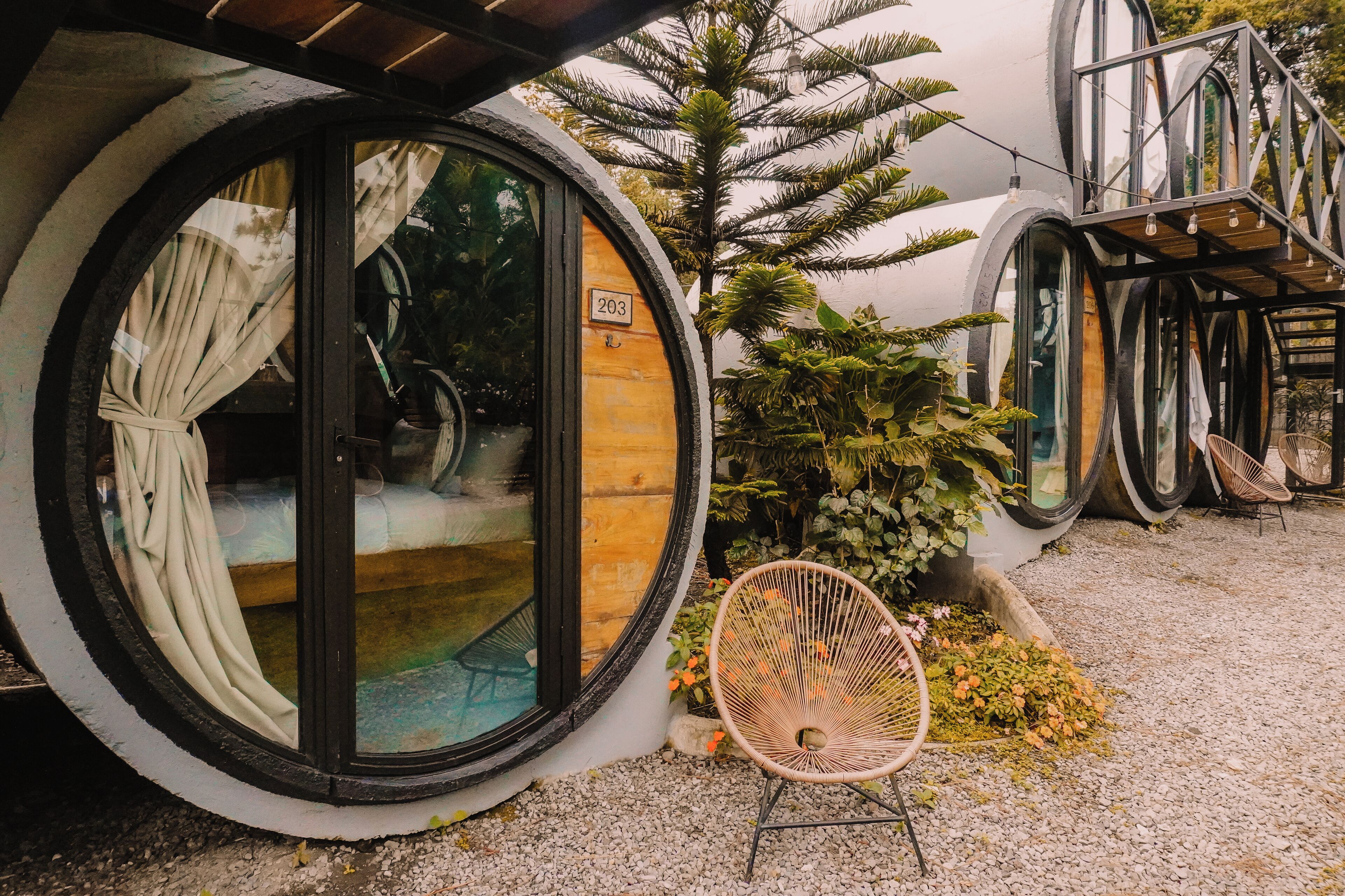 A unique spherical building with a wooden interior, a number '203' on the door, and a small garden area with a chair and plants.
