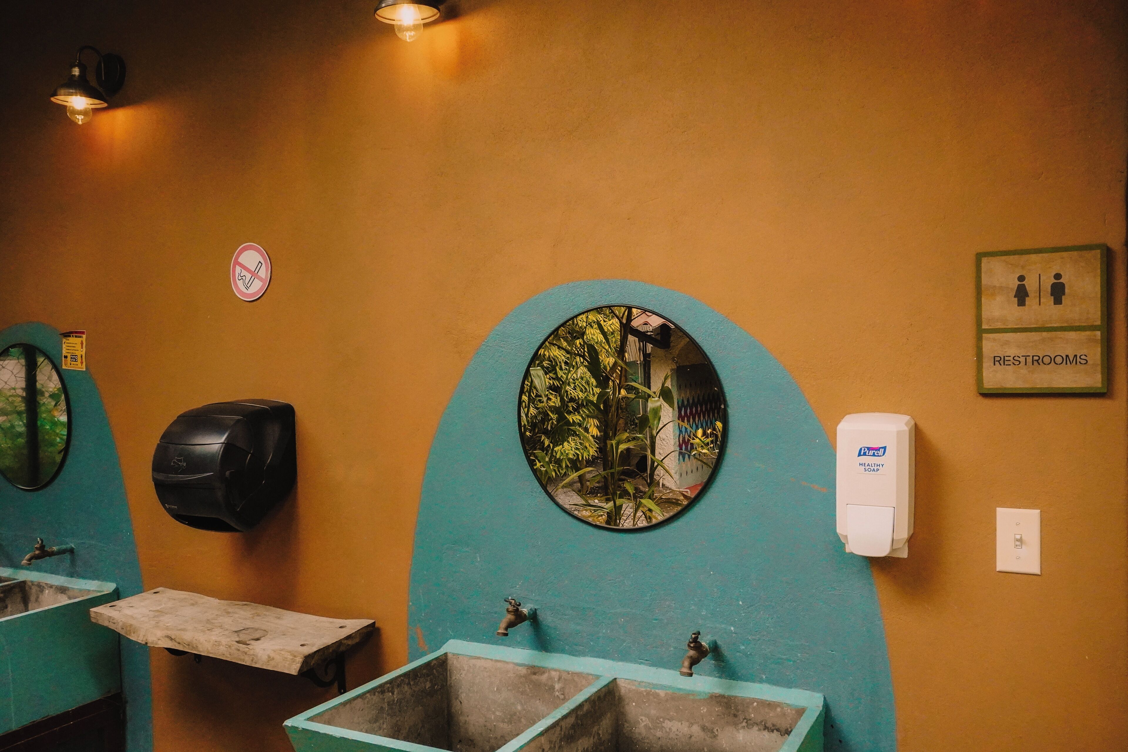 Baño con dos lavamanos, dispensador de jabón, cartel de prohibido fumar y un espejo que refleja árboles.