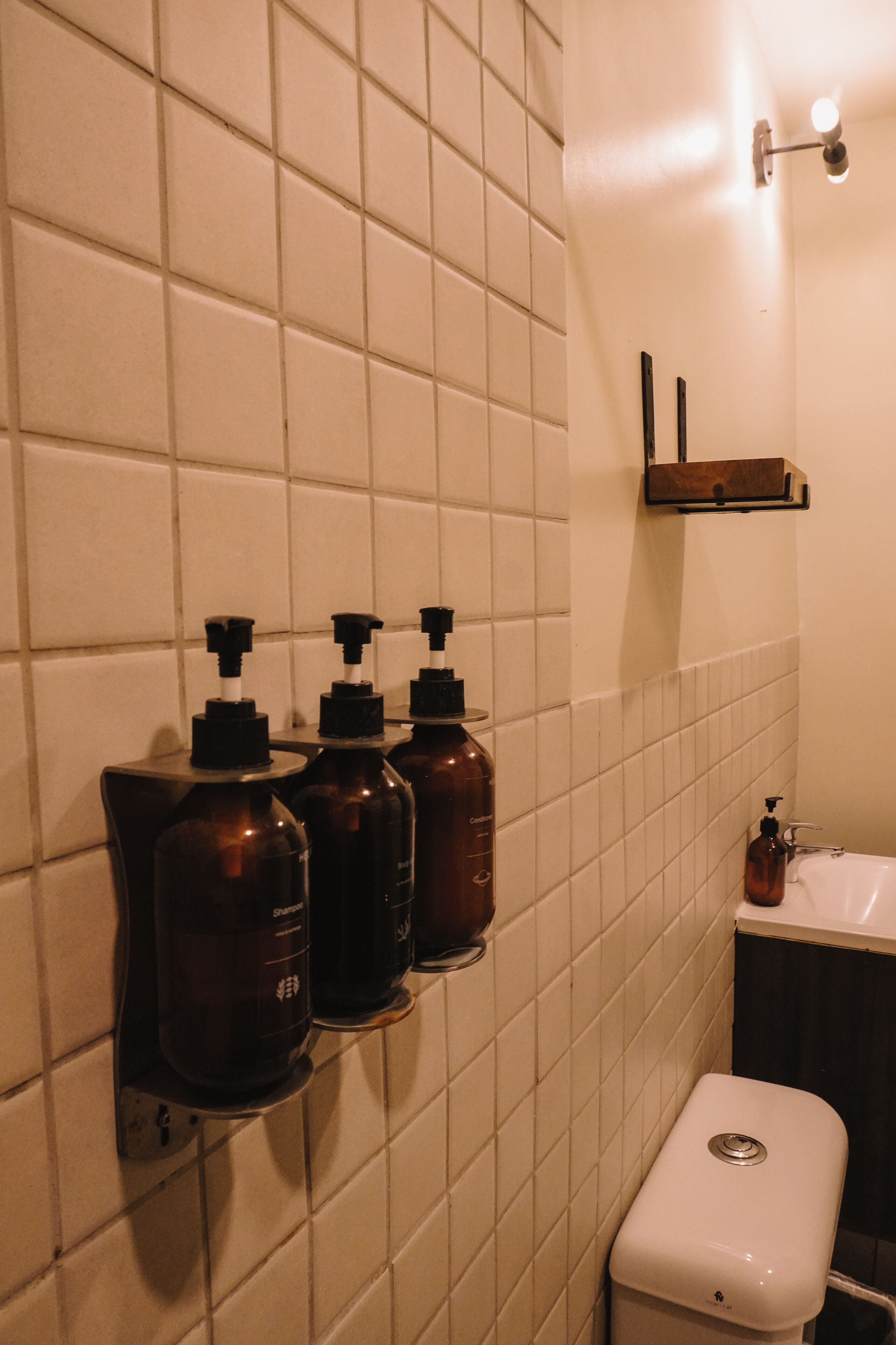 A bathroom with white tiled walls, a white toilet, and three brown dispensers on a wall-mounted shelf.
