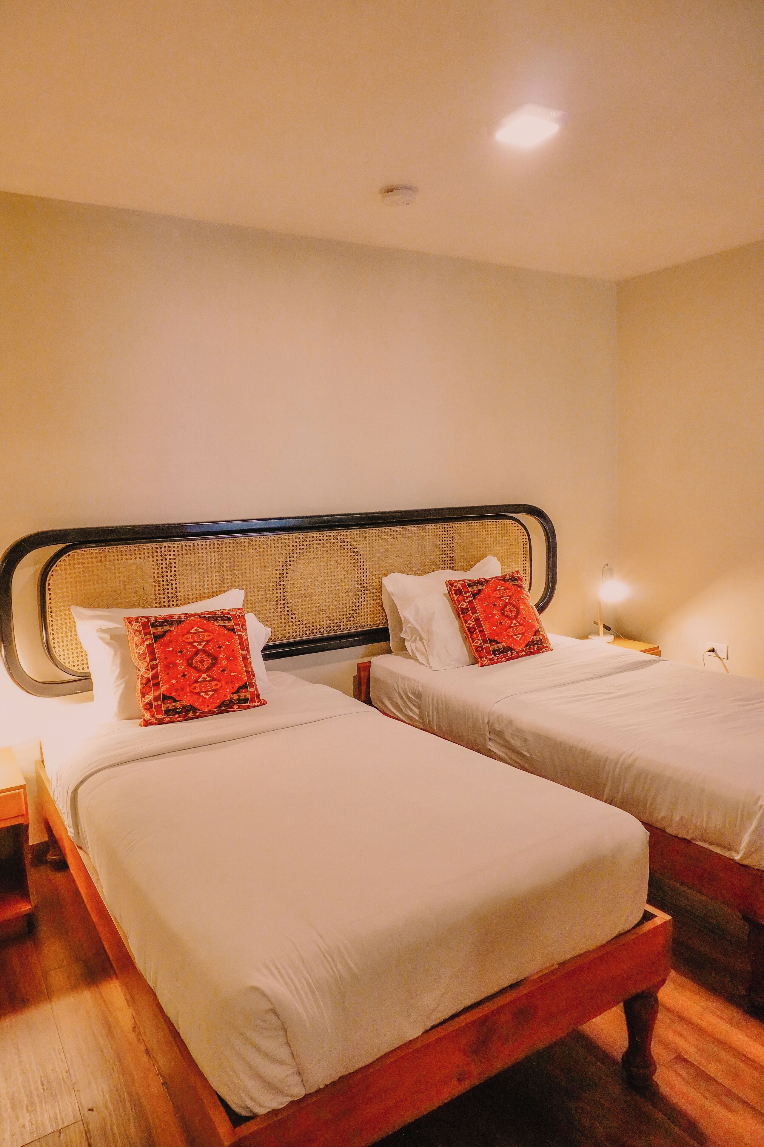 A hotel room with two single beds, each with white bedding and a red patterned pillow. The beds have wooden frames and a woven headboard. There is a nightstand with a lamp on each side of the beds.