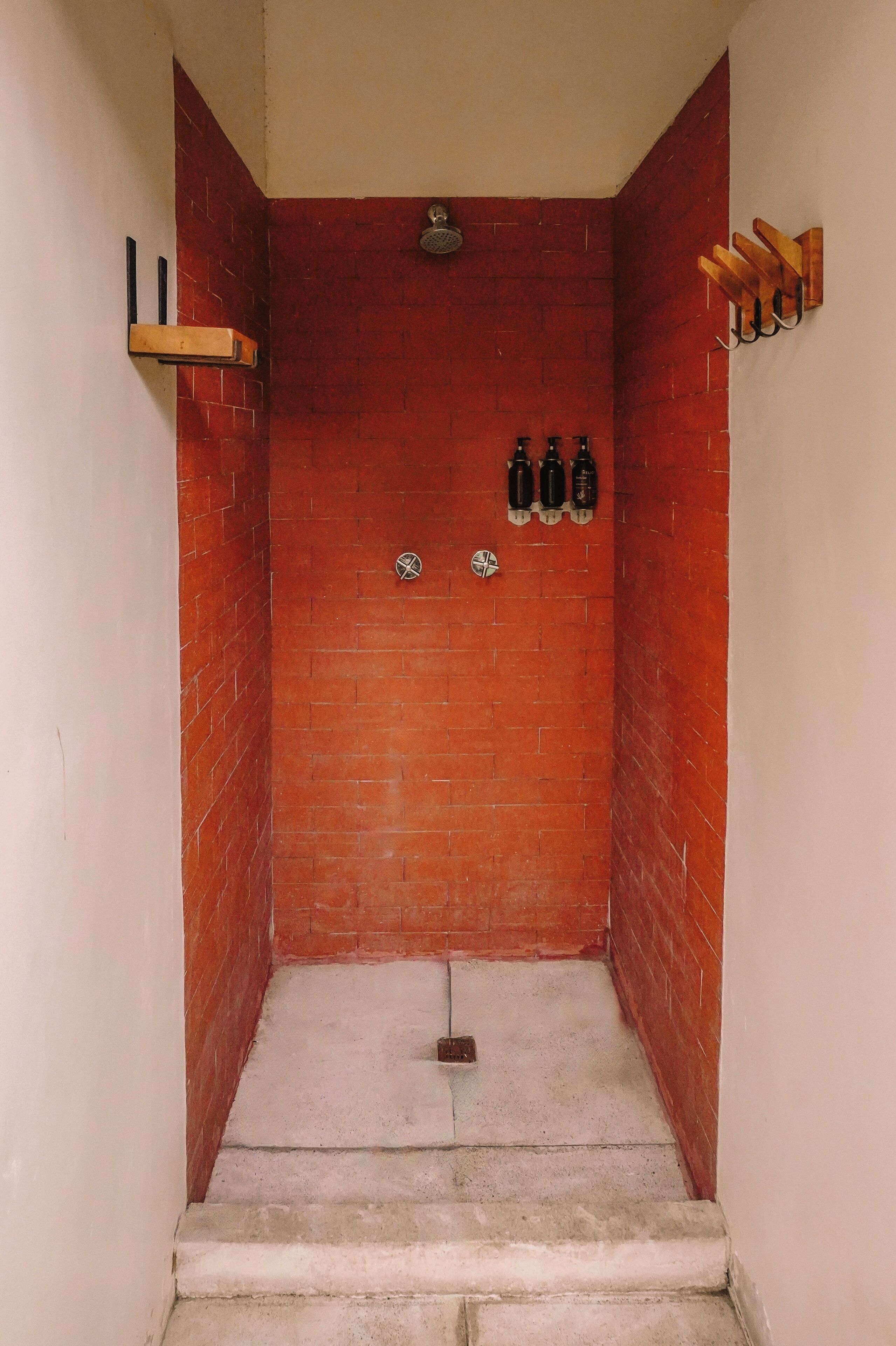 A narrow, enclosed shower area with red brick walls, a small shelf on the left, and a hook bar on the right.