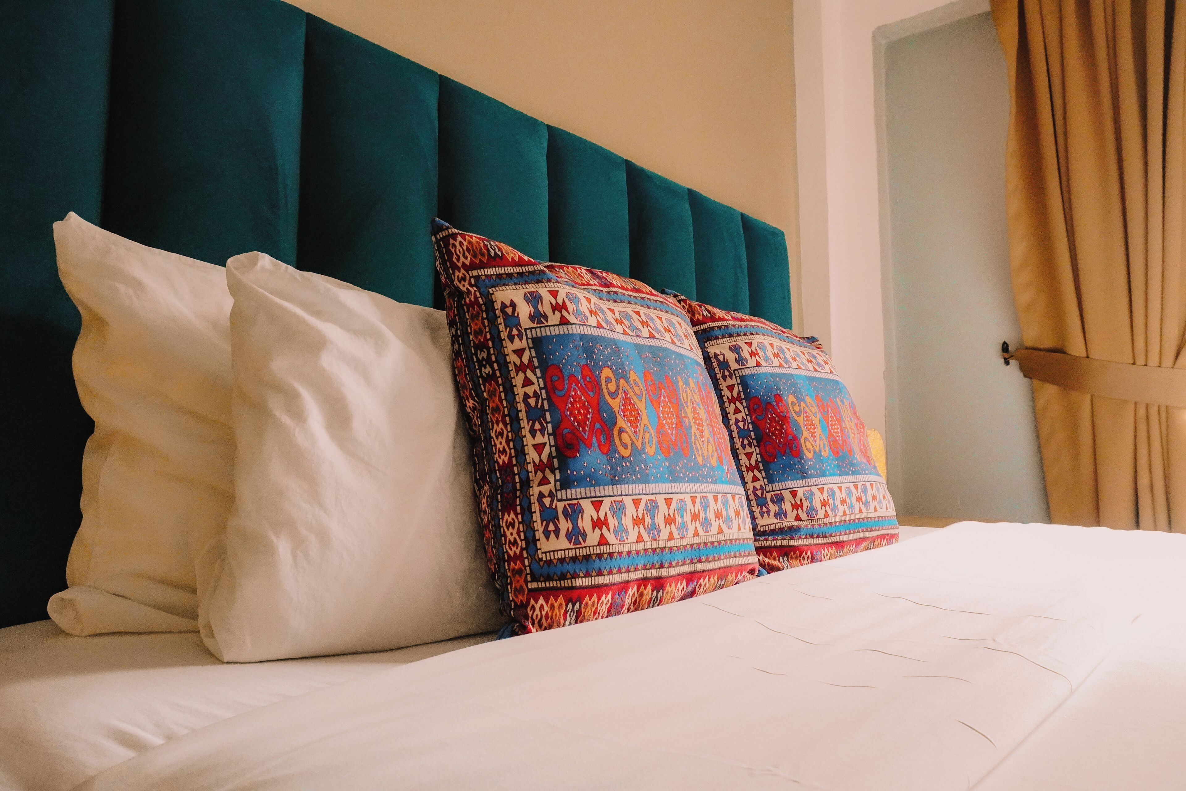 A bed with white and patterned pillows, teal headboard, and a beige curtain.