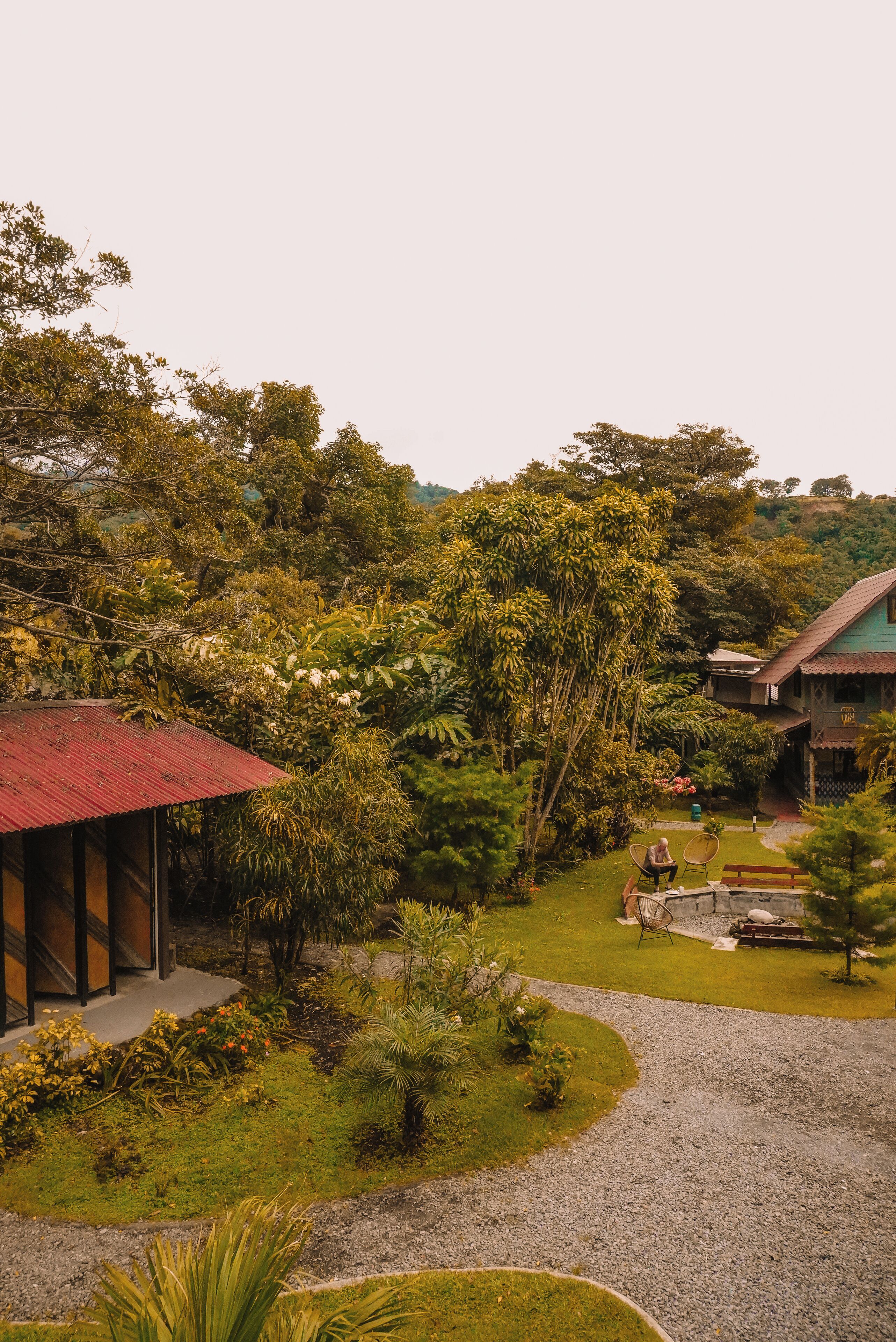 A lush garden with a red-roofed building, a gravel path, and a seating area with chairs.