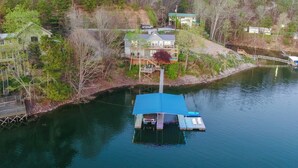 Una casa flotante amarrada a un río, con terraza y zona de estar, rodeada de árboles y un muelle con un pequeño edificio.