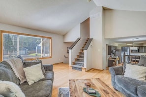 A spacious living area with a staircase leading to a kitchen, large windows, and a cozy seating arrangement.