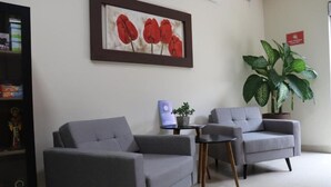Un área de recepción con dos sillones grises, una mesita, una planta en maceta y un cuadro de flores rojas en la pared.