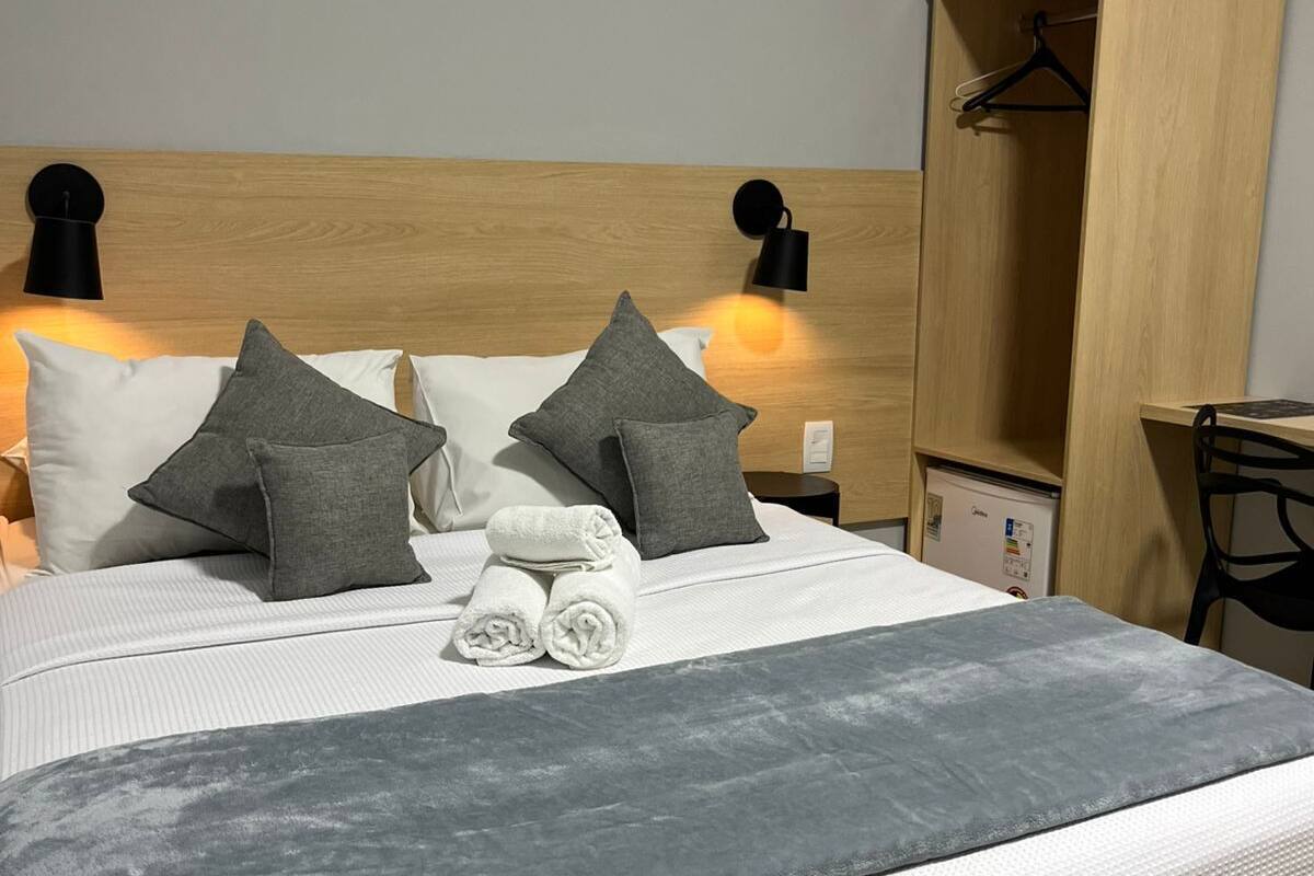 A neatly made bed with pillows, a grey blanket, and a rolled towel. There are two wall-mounted lamps on either side of the bed.