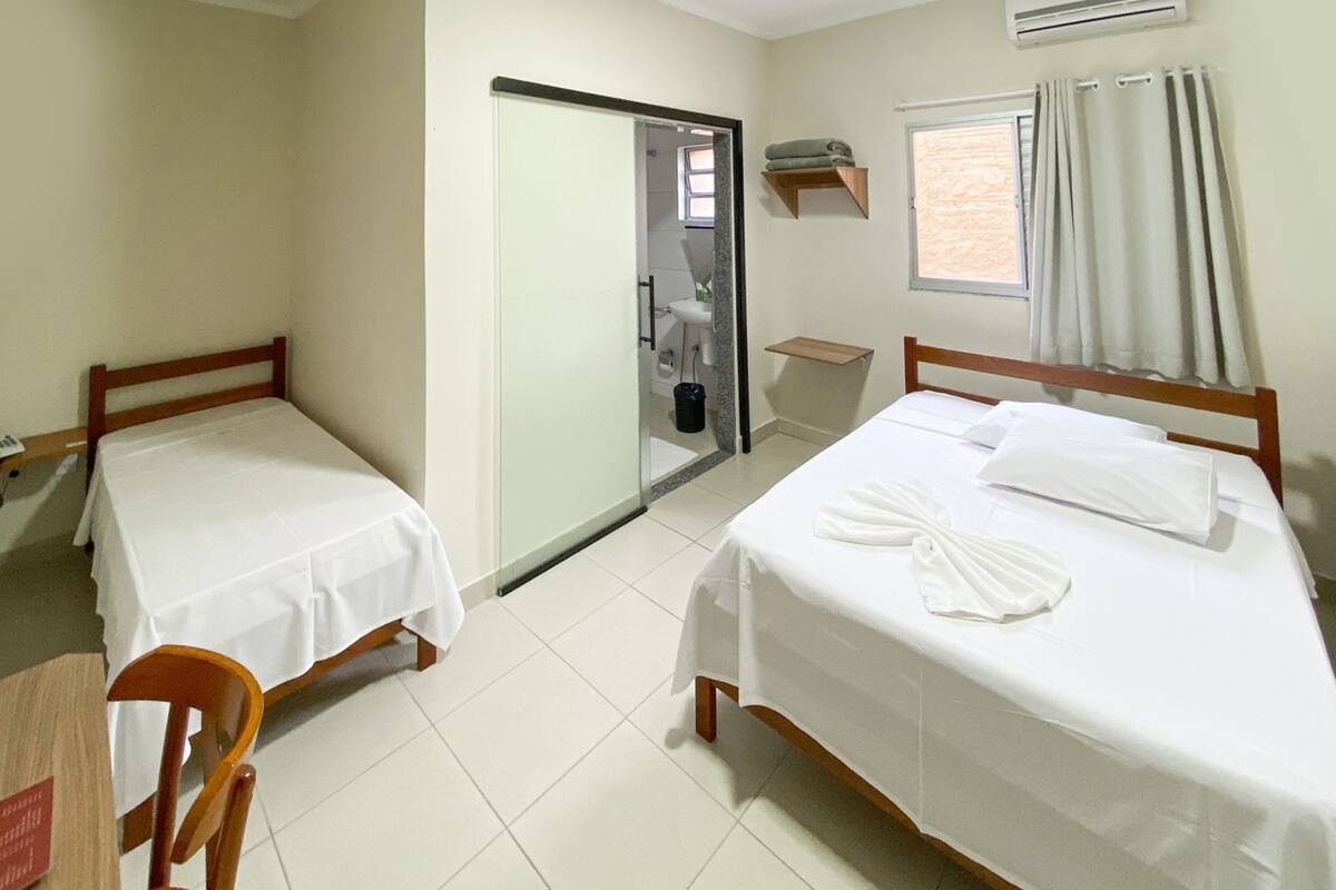 A hotel room with two single beds, a small table, a chair, a bathroom with a shower, and a window with curtains.