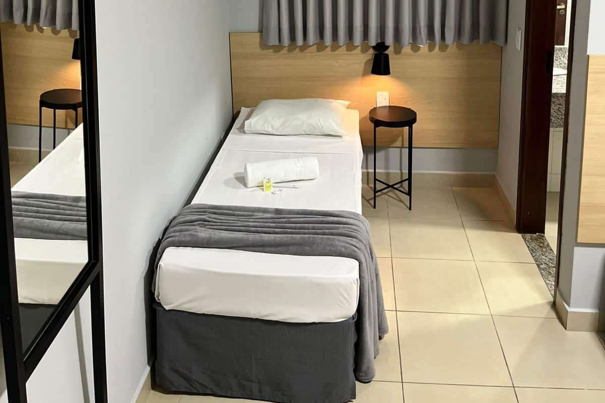 A compact hotel room with a single bed, a bedside table with a lamp, a small stool, and an air conditioning unit mounted on the wall.