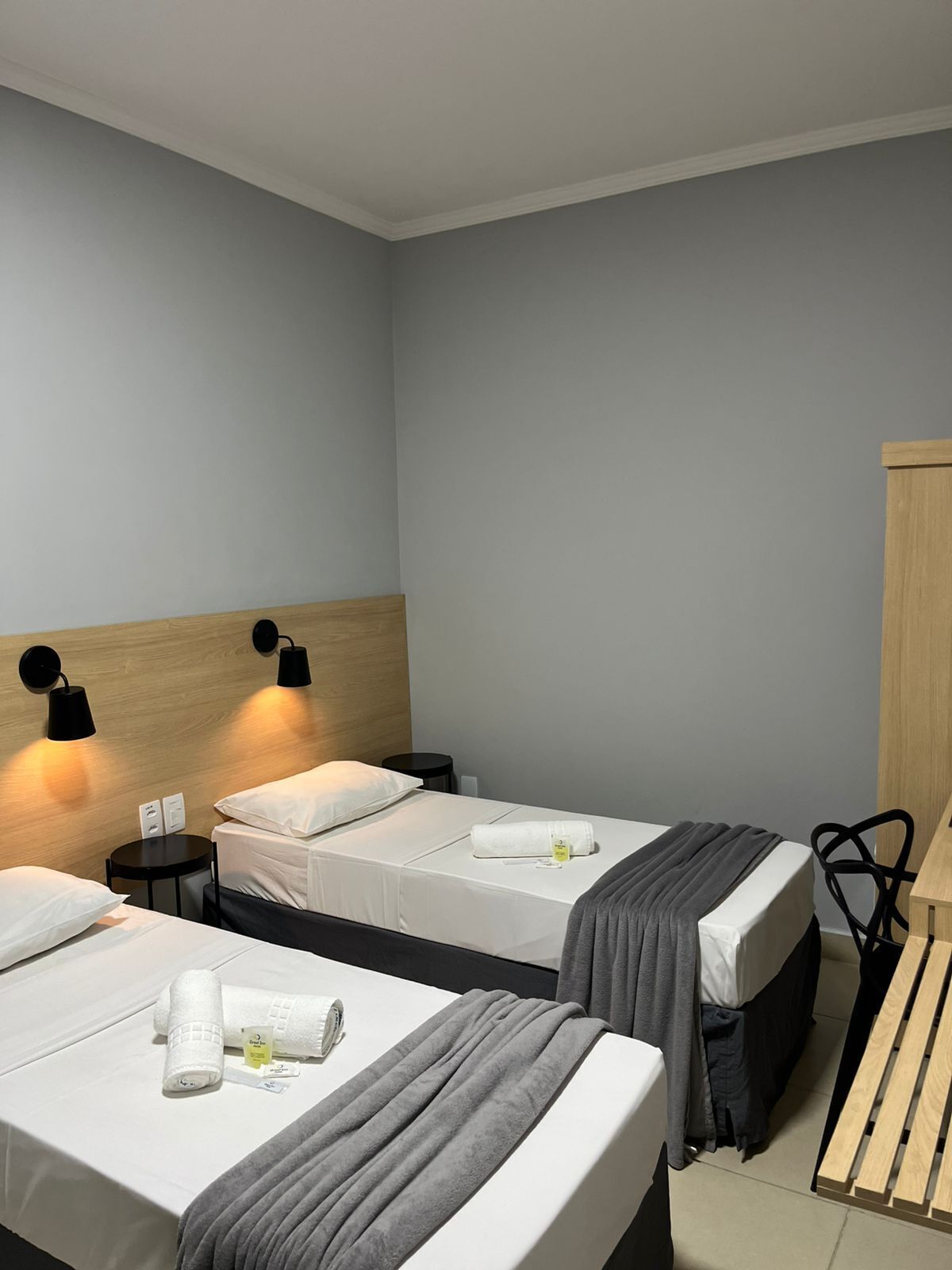 A hotel room with two beds, each with a wooden headboard, white linens, and a grey blanket. There are two black wall sconces providing light, a small round table with a chair, and a wooden bench.