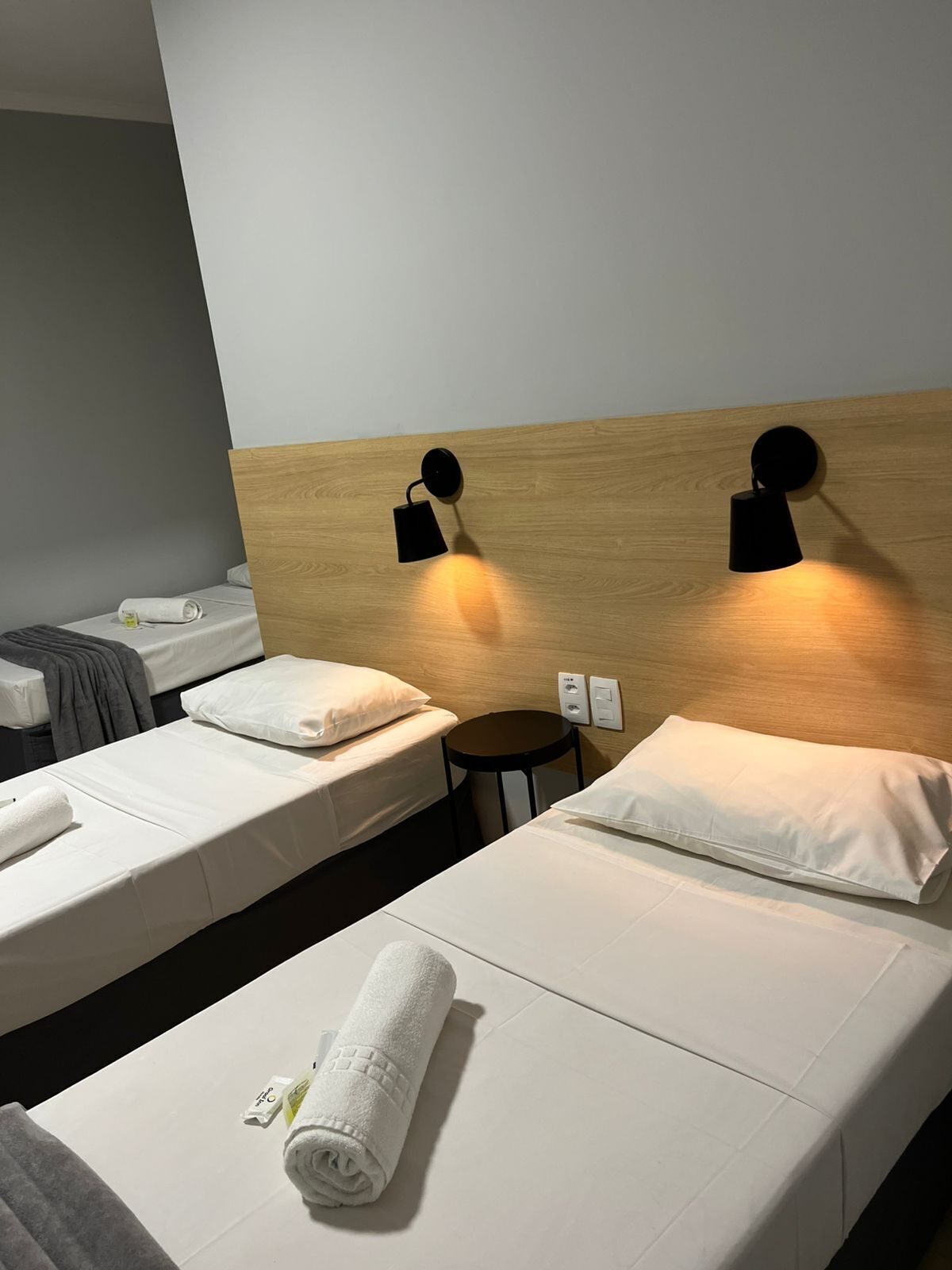 A hotel room with two single beds, each with white linens and pillows, a wooden headboard, and two wall-mounted black lamps.