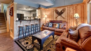 A cozy living room with a leather sofa, a coffee table, and a patterned rug. A kitchen with bar stools is visible in the background. - Ski In Ski Out Stagecoach Lift at Heavenly 3BR 3BA (Stateline)