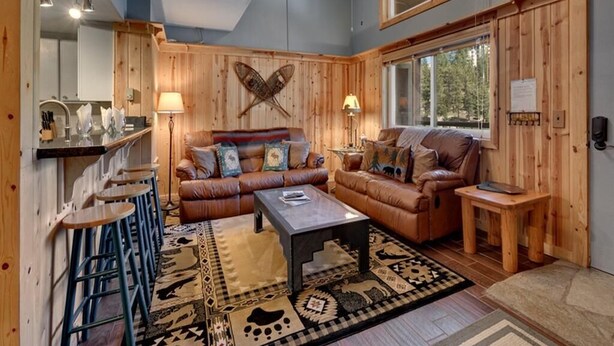 A cozy living room with a wooden ceiling, a ceiling fan, a large window, a sofa, a coffee table, and a patterned rug. - Ski In Ski Out Stagecoach Lift at Heavenly 3BR 3BA (Stateline)