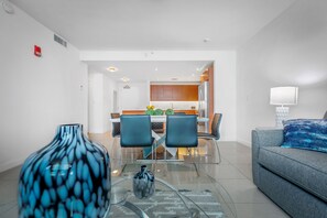A modern living room with a glass coffee table, two blue chairs, a grey sofa, and a dining area in the background.