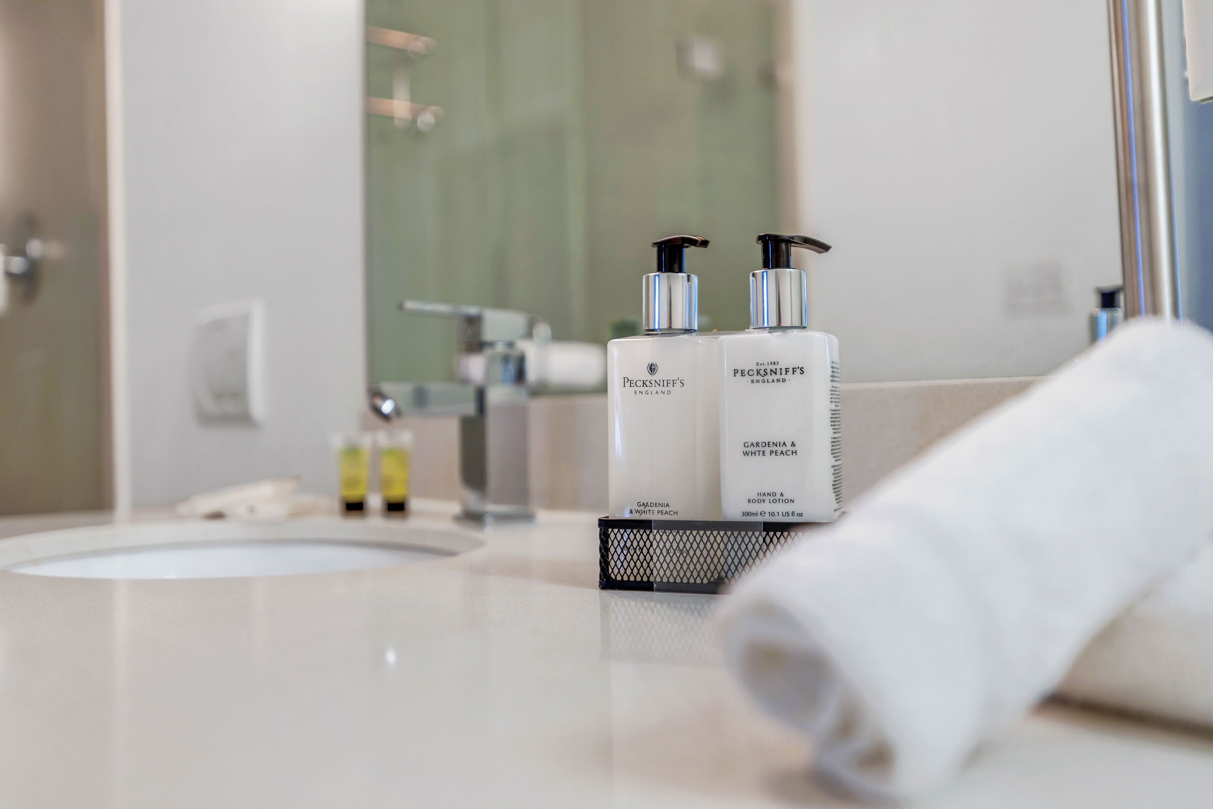 Une salle de bain avec un lavabo blanc, deux flacons de lotion pour le corps de la marque Pecksniffs et une serviette posée sur le comptoir.