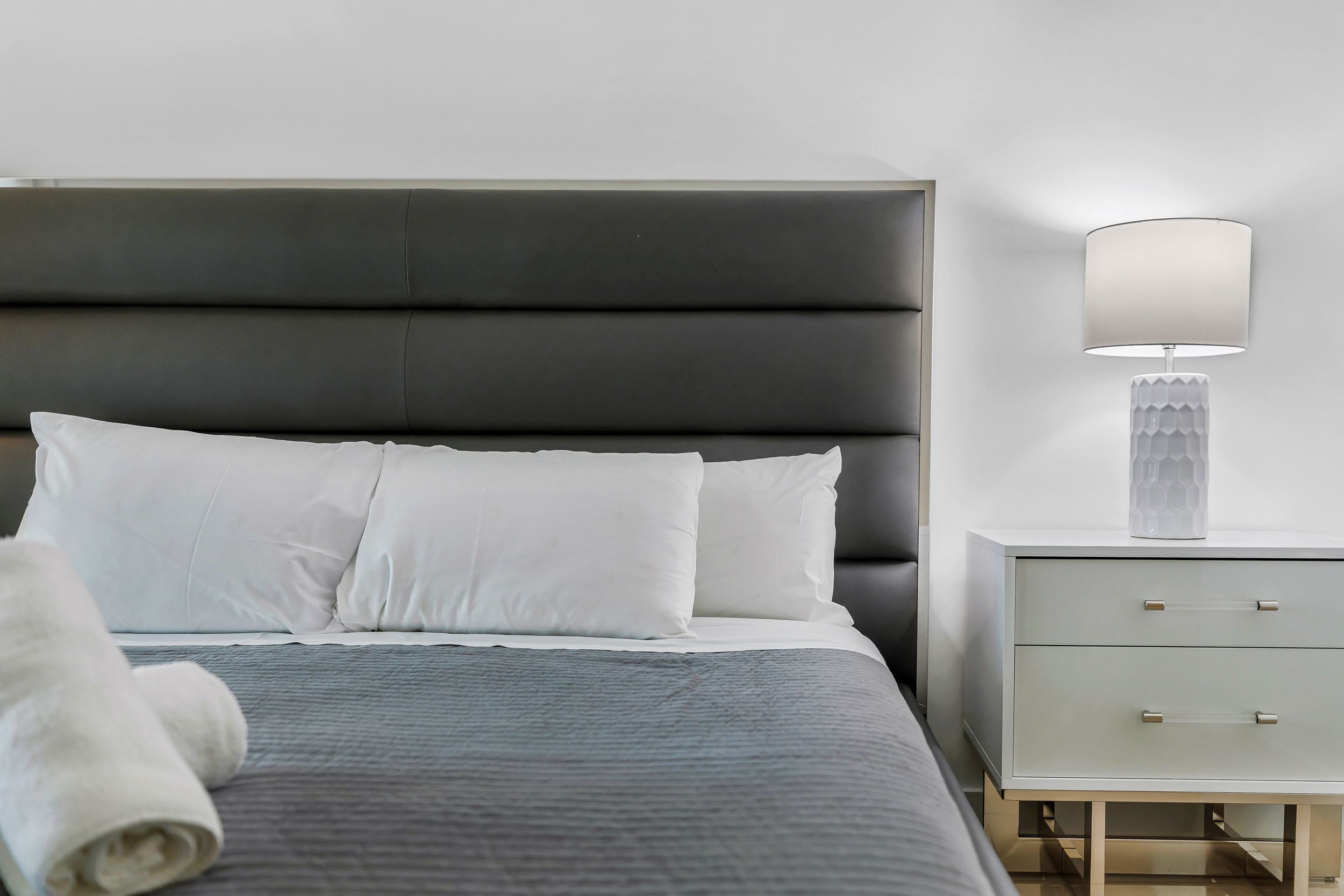 A neatly made bed with white linens, a grey bedspread, and a rolled white towel on the side.