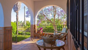 Eine überdachte Terrasse im Freien mit Korbmöbeln, einem Glastisch und Blick auf Grünpflanzen und violette Blumen.