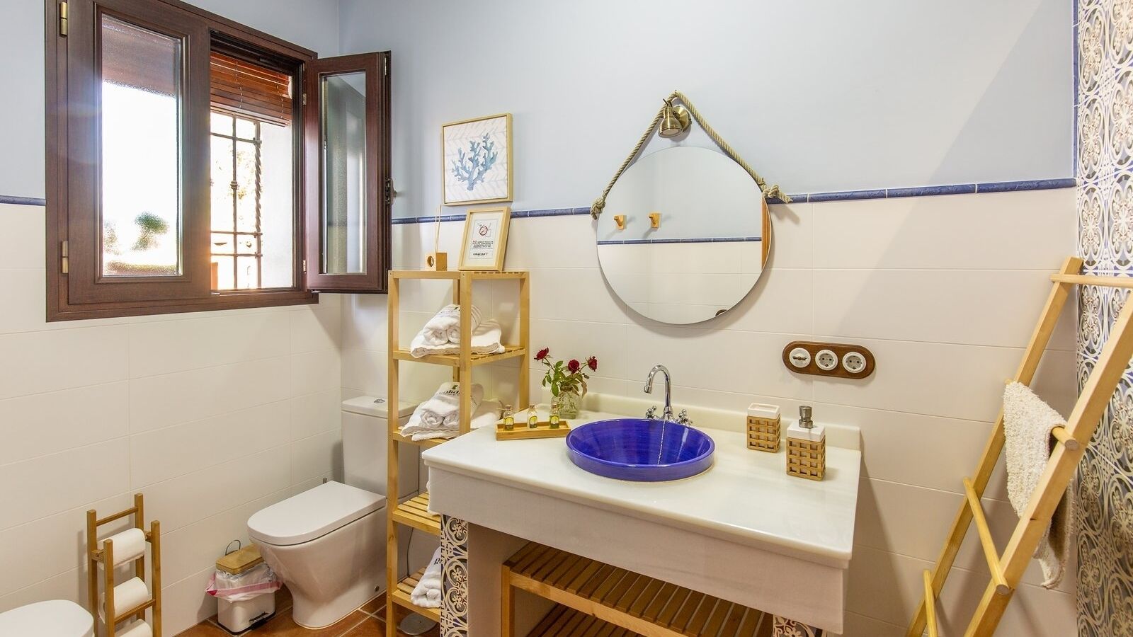 A bathroom with a toilet, a sink with a blue basin, a wooden shelf with towels, a mirror, and a window with blinds.