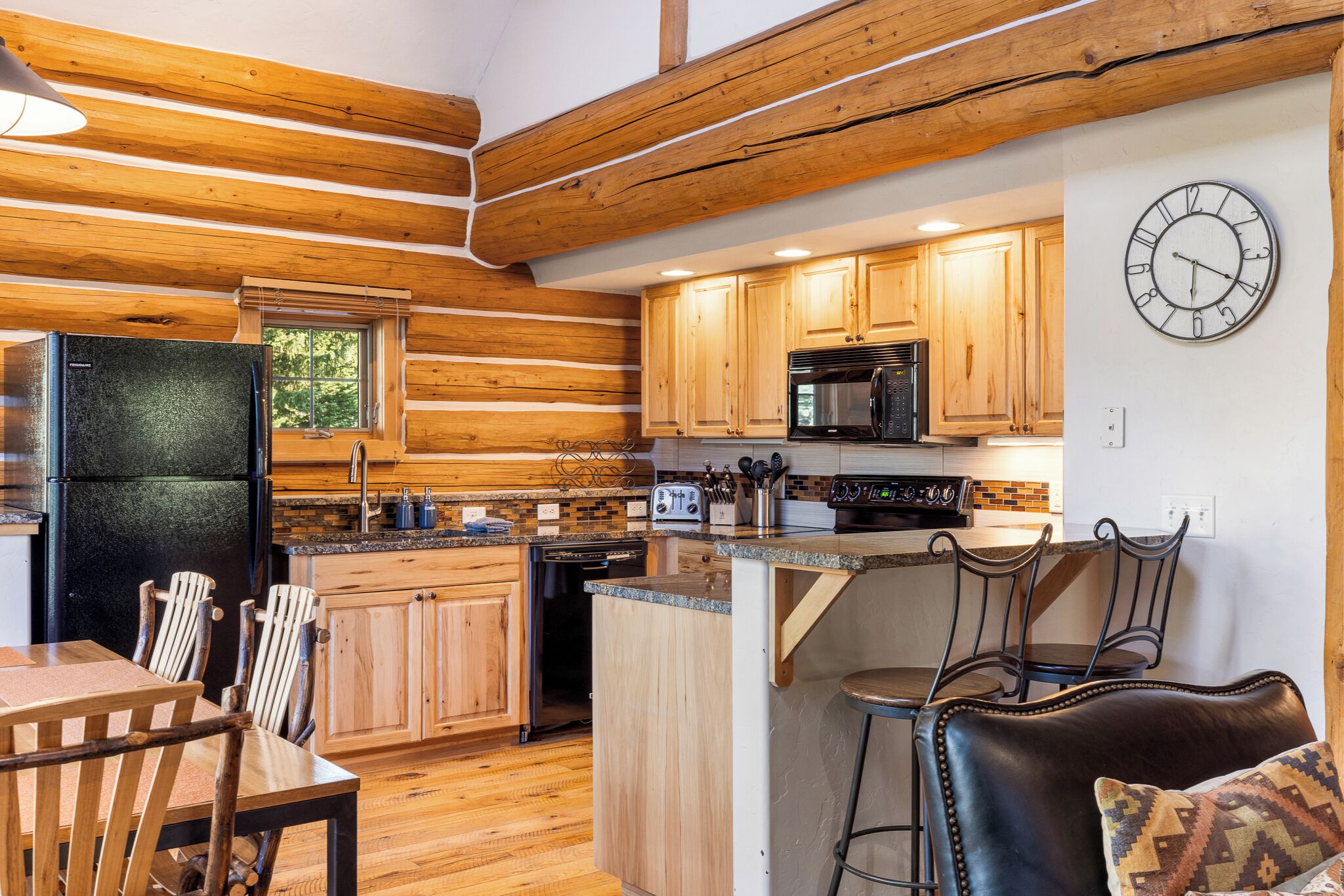 A log cabin-style kitchen with wooden cabinets, a black refrigerator, a microwave, and a clock on the wall.
