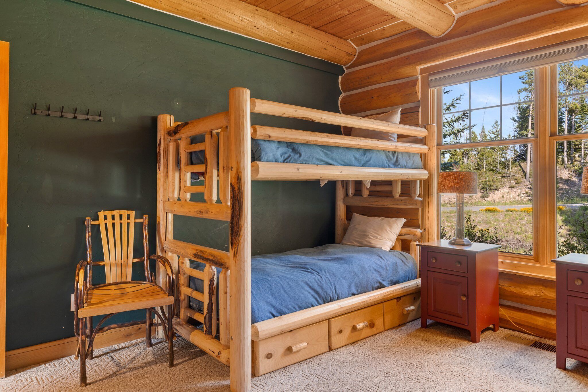 Una cama individual con estructura de madera, una silla, una mesita de noche y una ventana que da a vistas de árboles.
