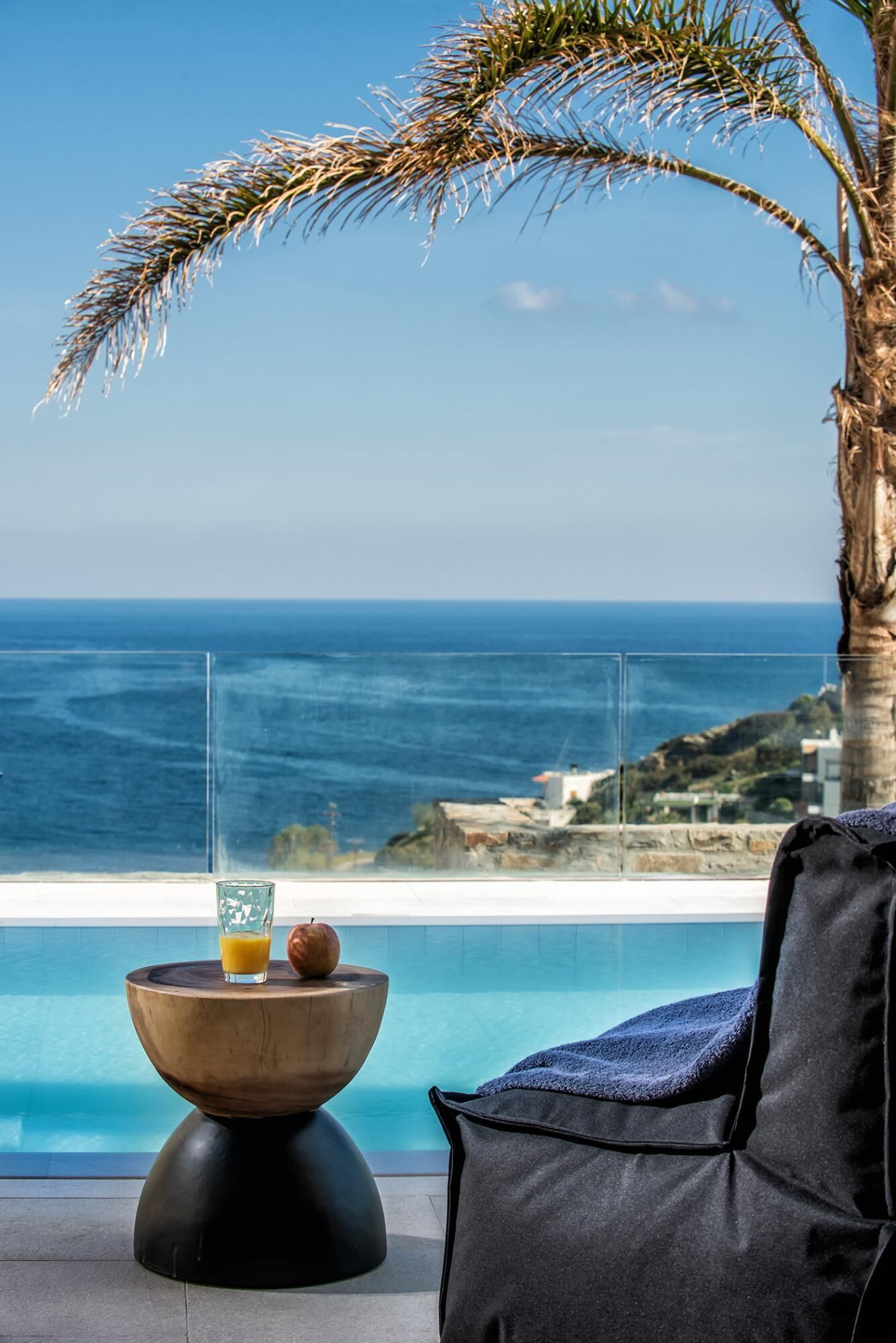 A poolside area with a view of the ocean, a palm tree, a glass table with an apple and a glass of orange juice, and a black lounge chair.