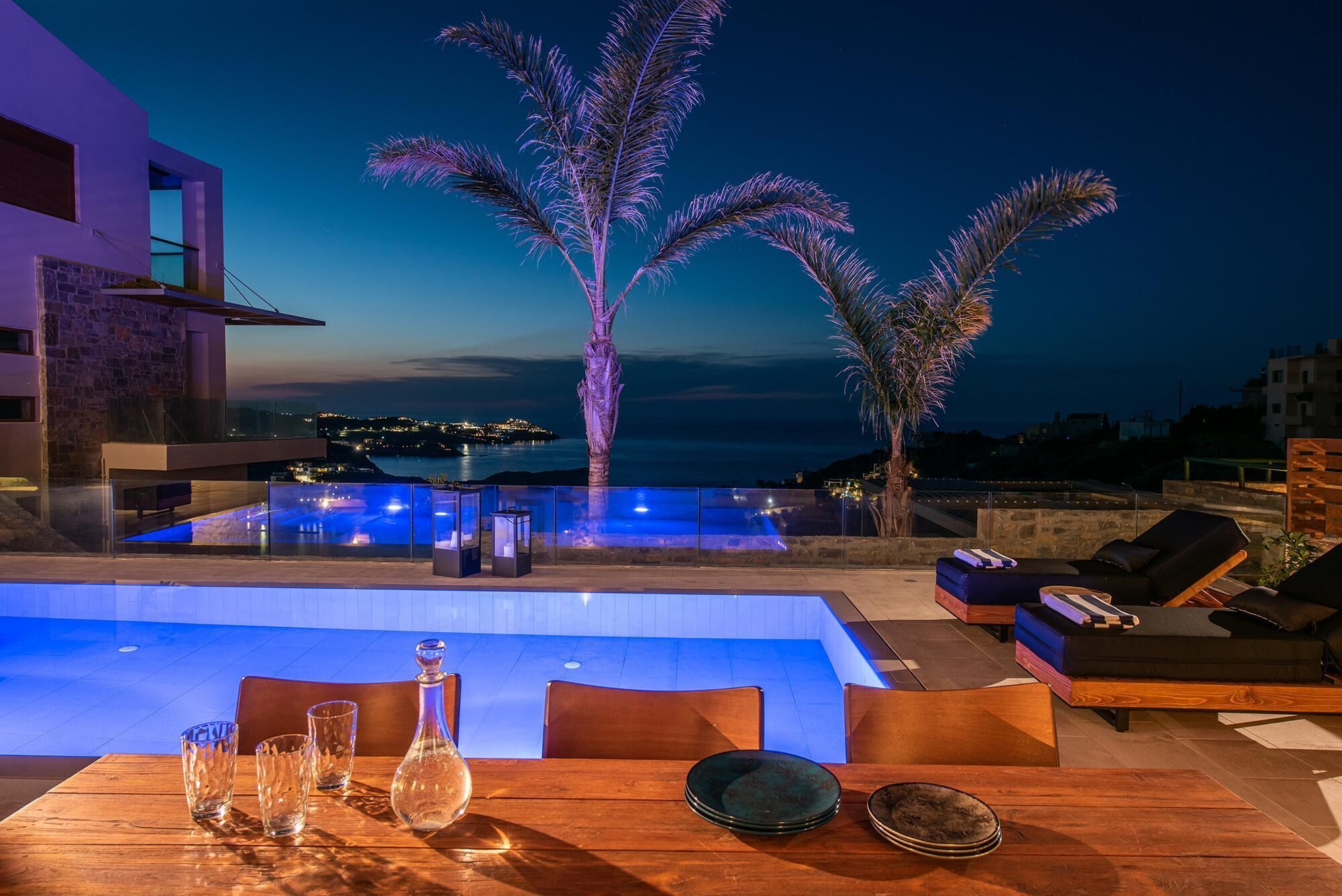 Un espace piscine aménagé sur le toit, avec des chaises longues et une table à manger, offrant une vue sur le paysage urbain côtier au crépuscule.