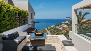 A balcony with wicker furniture, a table with fruit, and a view of the sea and hillside buildings. - Alpha House in Crete (Malevizi)