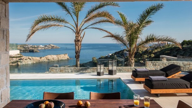 A poolside area with a view of the sea, palm trees, and a stone wall. - Alpha House in Crete (Malevizi)