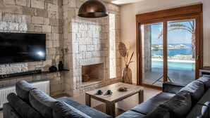 A living room with a stone wall, a fireplace, a television, and a view of the sea. - Alpha House in Crete (Malevizi)