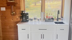 Ett kök med vita skåp, en kaffestation och ett stort fönster med utsikt över träd.