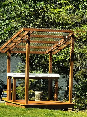 A wooden pergola structure with a table underneath, situated in a grassy area with a body of water and dense foliage in the background.
