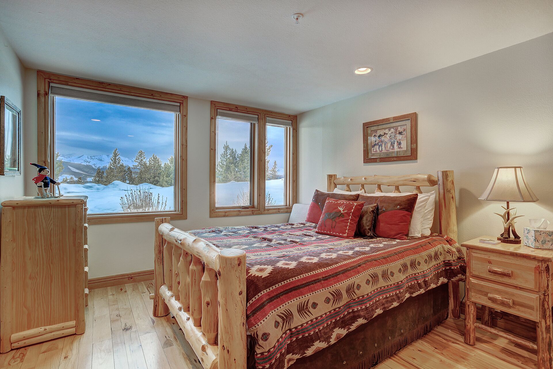 Quarto com cama de madeira, duas janelas com vista para uma paisagem nevada, mesa de cabeceira com abajur e um quadro na parede.