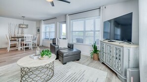 Una sala moderna con ventilador de techo, televisor, comedor con mesa y sillas, y una zona de estar con sofá y reposapiés.