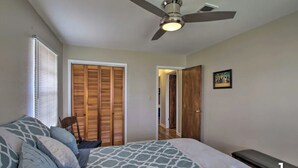 Quarto com cama, ventoinha no teto, janela com persianas, porta de madeira e quadro emoldurado na parede.