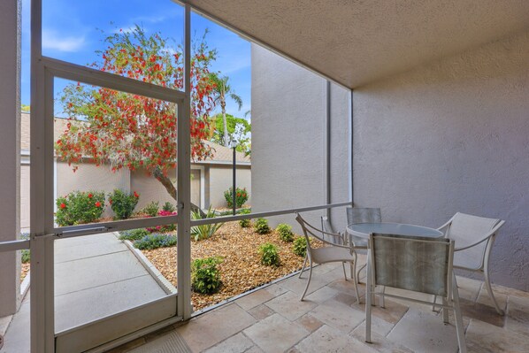 An outdoor patio with a table and chairs, a view of a garden with plants and a tree, and a building in the background.