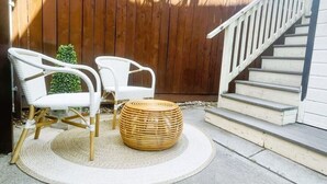 Eine Terrasse mit zwei weißen Korbstühlen, einem runden gewebten Hocker und einem kleinen Topfpflanzen. Im Hintergrund sind ein Holenzaun und eine weiße Treppe zu sehen.