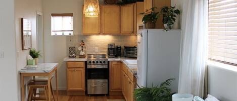 A compact kitchen with wooden cabinets, a stove, and a refrigerator. There is a dining area with a table and chairs, and a window with blinds.
