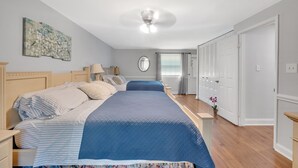 Un dormitorio con cama, mesitas de noche, ventilador de techo, ventana con cortinas y puerta con espejo.