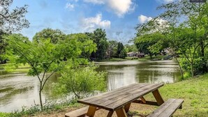Una mesa de picnic junto a un lago, con vegetación circundante y casas al fondo.