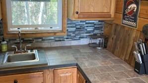 A kitchen with wooden cabinets, a stone backsplash, a window, and a sign advertising salmon. - Cozy Quiet Alaskan Cabin (Soldotna)