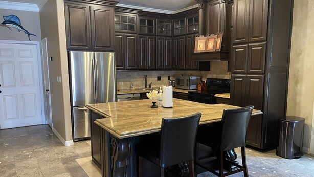 Una cocina con armarios de madera oscura, electrodomésticos de acero inoxidable, encimera de mármol y sillas negras.