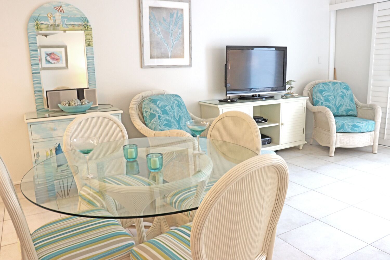 Un salon avec une table en verre, des fauteuils en osier, un téléviseur et un meuble blanc.