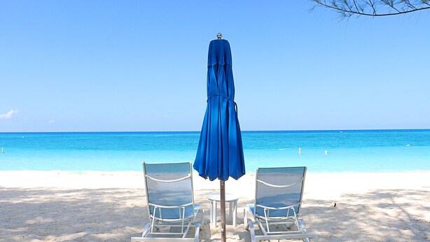 A beach with two white lounge chairs and a blue umbrella.