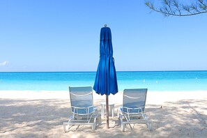 A beach with two white lounge chairs and a blue umbrella. - Galleon Villas by Hello Cayman Vacation (Seven Mile Beach)